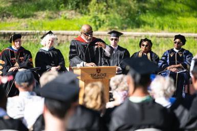 A Black man in an academic robe gestures over a podium toward an audience wearing black robes and graduation caps. 在他身后是五个人，也穿着学术袍.