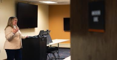 Dr. Jen Bossard speaks at the front of the College of Business's technology classroom, 位于林肯的弗雷德·布朗中心. 一扇打开的门部分遮住了图像, 博萨尔身后是两个大电视屏幕, 一个平板电脑和一个科技柜.